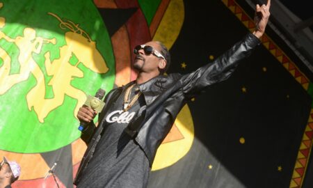 Male rapper on stage wearing sunglasses and a black leather jacket, raising one arm as he holds a microphone.