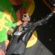 Male rapper on stage wearing sunglasses and a black leather jacket, raising one arm as he holds a microphone.
