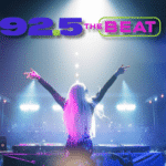 DJ on stage with arms raised performing to a large crowd at a concert, 92.5 THE BEAT logo glowing above the scene