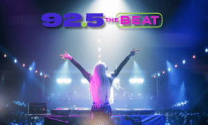 DJ on stage with arms raised performing to a large crowd at a concert, 92.5 THE BEAT logo glowing above the scene