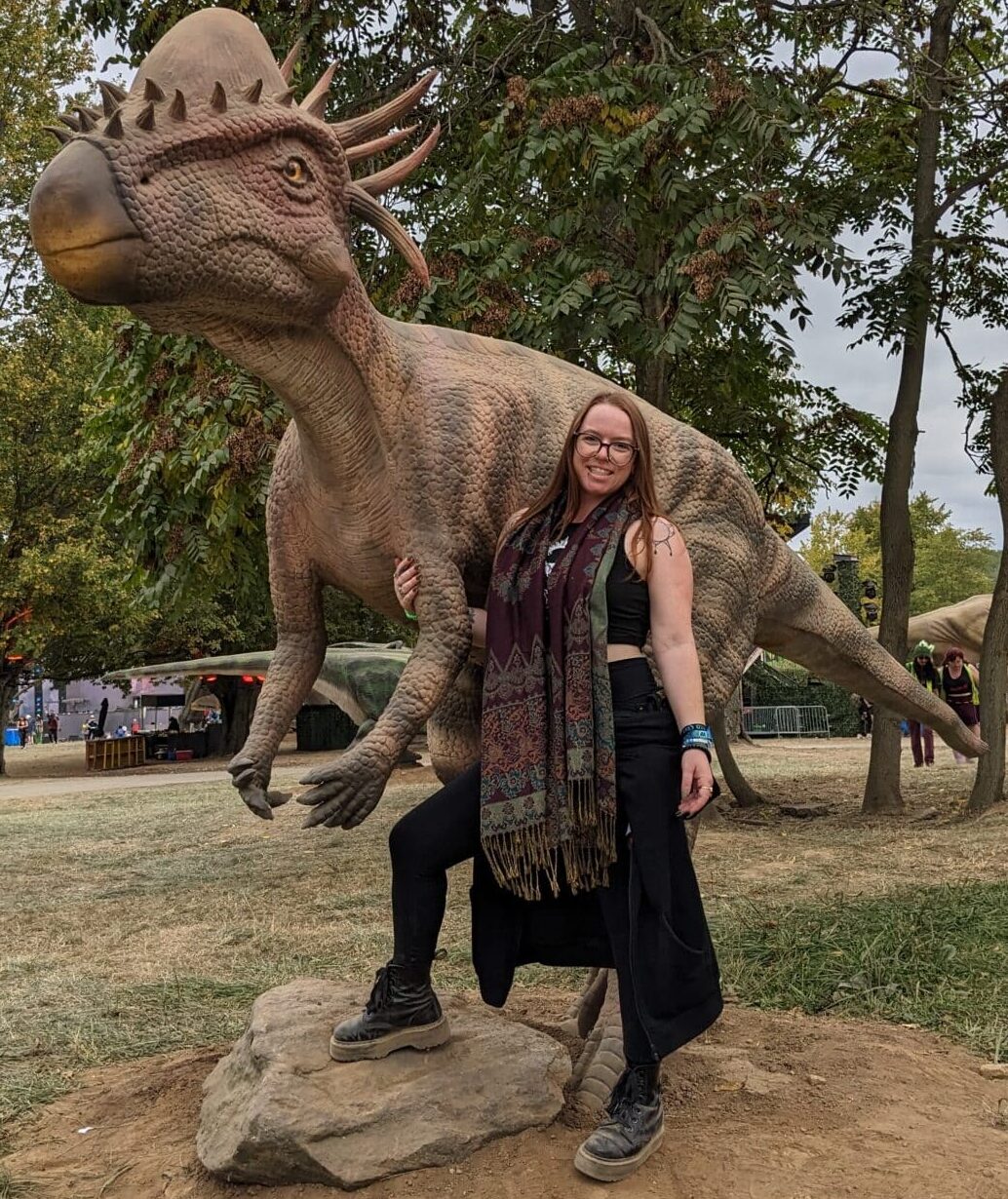 Caity and Packi | 92.5 The Beat Person posing with a large dinosaur statue in a park, woman leaning against the dinosaur's leg and smiling under trees.",