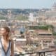 Woman in a silver dress stands on a balcony, with a blurred city skyline and a domed building in the distance behind her.