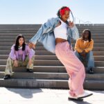 Three friends sit and pose on outdoor concrete steps; center person dances in pink pants, white top, denim jacket, and red headband.