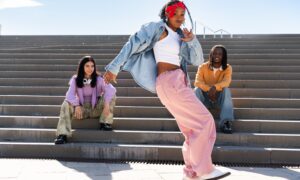 Three friends sit and pose on outdoor concrete steps; center person dances in pink pants, white top, denim jacket, and red headband.