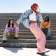 Three friends sit and pose on outdoor concrete steps; center person dances in pink pants, white top, denim jacket, and red headband.